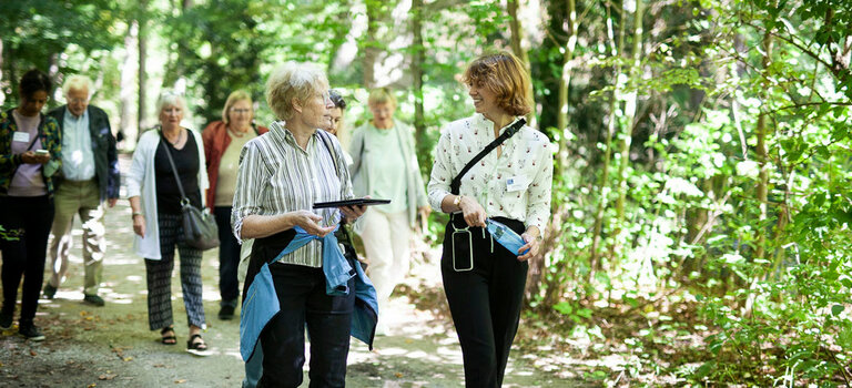Gruppe von Senior*innen und Projektmitarbeiterinnen von "Digital mobil im Alter" spazieren fröhlich durch den Englischen Garten in München. Sie halten Tablets und Smartphones in der Hand und unterhalten sich angeregt.