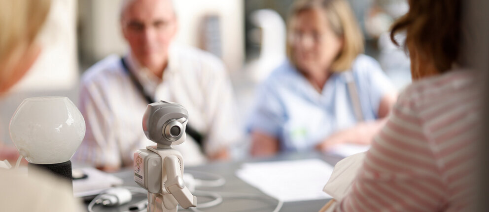 Auf dem Tisch steht ein kleiner Roboter, im Hintergrund sind verschwommen ältere Personen zu sehen