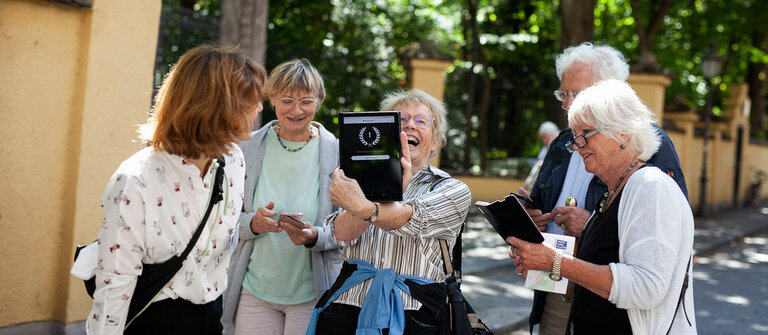 Eine Gruppe von Senior*innen mit Unterstützerin beim Digitalen Spaziergang. In der Mitte hält die Siegerin ein Tablet hoch, auf dem eine 1 für erster Platz zu sehen ist