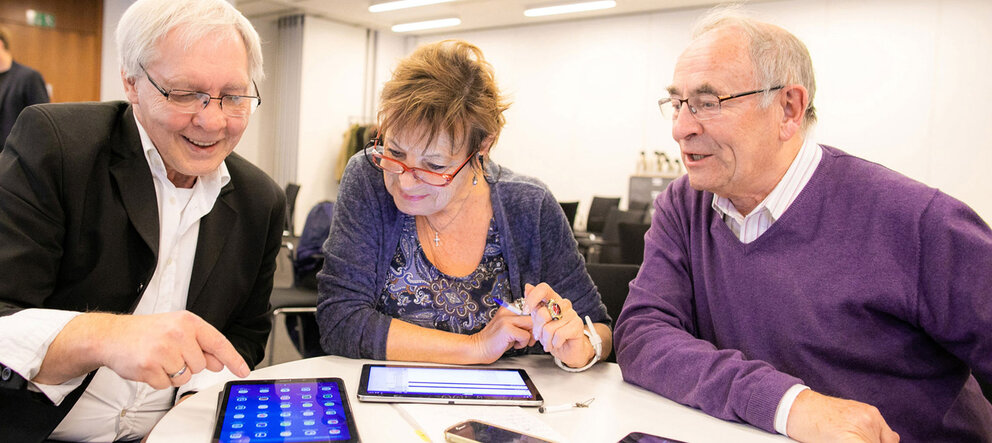 Zwei ältere Herren und eine ältere Dame sitzen an einem Tisch und bedienen Tablets